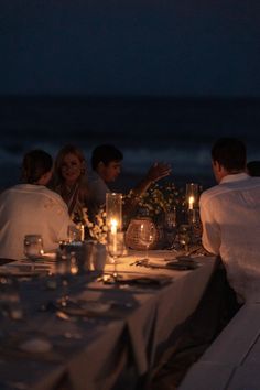 a group of people sitting at a table with candles in front of them and the ocean behind them