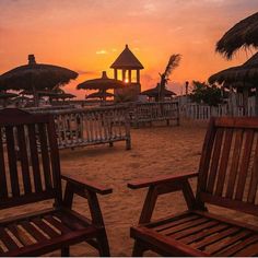 two wooden chairs sitting on top of a sandy beach next to umbrella covered buildings at sunset