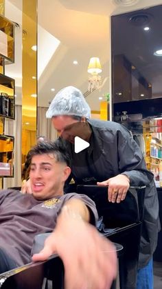 a man getting his hair cut by a barber in a store with another man pointing at him