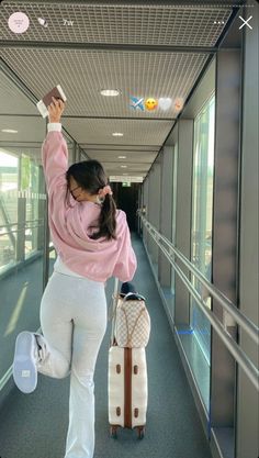 a woman in white pants and pink sweater pulling luggage down a hall way with her hand up