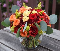 a glass vase filled with red, orange and yellow flowers on top of a wooden table