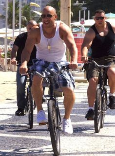 two men riding bikes down the street in front of other people wearing black vests