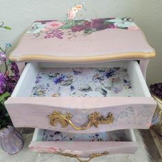 an ornate pink and gold jewelry box with floral designs on it, sitting next to purple flowers