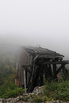 an old train trestle in the fog on a bridge over some rocks and grass