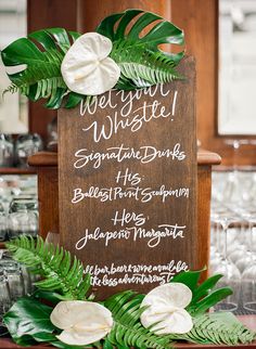 a wooden sign with white flowers and greenery on it sitting in front of wine glasses