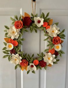 a wreath made out of paper flowers hangs on the front door with an orange, white and red flower arrangement