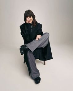 a woman sitting on top of a chair in a black coat and grey striped pants