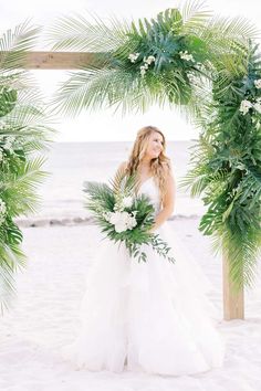 a woman in a white wedding dress standing under a palm tree arch on the beach