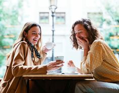 two women sitting at a table laughing and drinking coffee