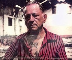 an old man with tattoos on his chest standing in front of a building and looking at the camera