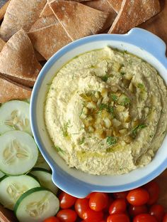 a bowl of hummus surrounded by sliced cucumbers, tomatoes and pita chips