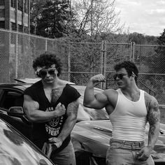 two men standing next to each other in front of a car with their fists raised