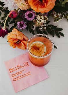 an orange drink sitting on top of a white table next to flowers and a pink napkin