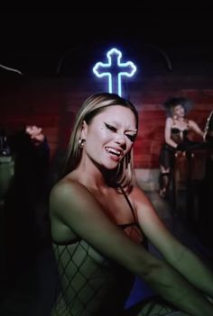 a woman sitting in front of a neon cross sign