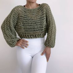 a woman wearing white pants and a green crochet sweater stands in front of a white wall