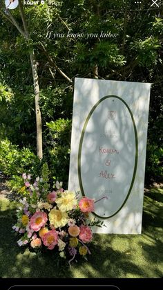 a sign that is in the grass with flowers around it and some trees behind it