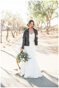 a woman in a white dress and black jacket holding a bouquet with trees in the background