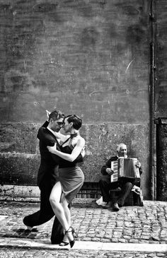 black and white photograph of two people dancing in front of an old brick wall, one man is playing the accordion