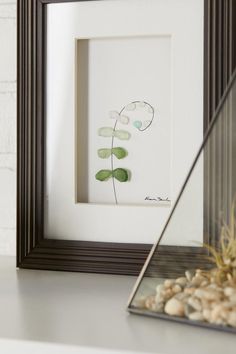 a white frame with some green leaves in it next to rocks and a potted plant