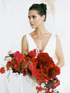 a woman in a white dress holding a bouquet of red flowers and greenery with her hands behind her back