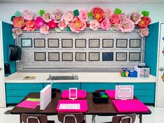 a table with pink and blue chairs in front of a wall decorated with paper flowers