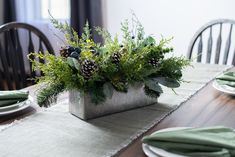 the table is set with pine cones and greenery in a silver container, along with green napkins