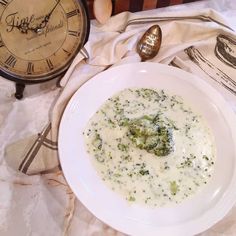 a bowl of broccoli and cheese soup next to an alarm clock on a table