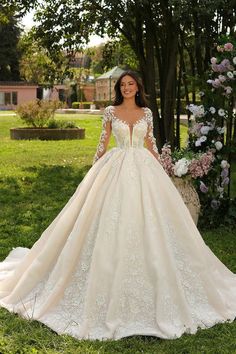 a woman wearing a wedding dress standing in the grass with trees and flowers behind her