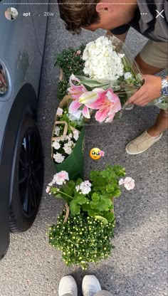 a man smelling flowers in front of a car with his feet propped up on the ground