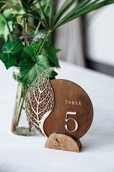 a wooden table number sitting next to a vase filled with green leaves and greenery