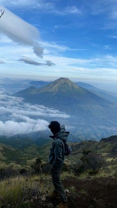 a man standing on top of a lush green hillside next to a mountain covered in clouds