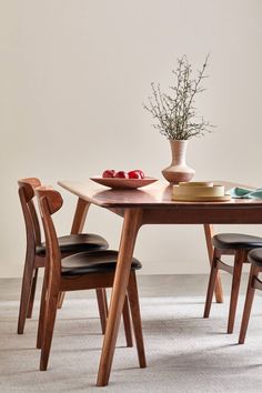 a wooden table topped with a bowl of fruit next to two chairs and a vase filled with flowers