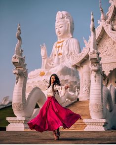 a woman in a long red skirt is dancing near a white statue with buddha statues behind her