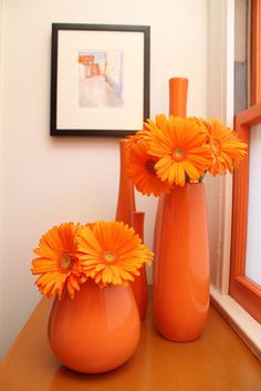 three orange vases with flowers in them sitting on a table next to a window