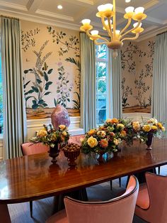 a dining room table with flowers and vases on it in front of two large windows