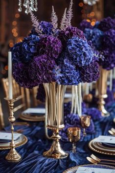 purple and blue flowers in gold vases on a table with silver plates, candles and place settings