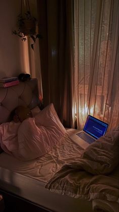 a laptop computer sitting on top of a bed next to a window with sheer curtains