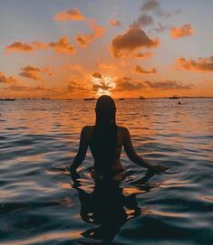 a woman is sitting in the water with her back turned to the camera as the sun sets