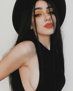 a woman with long black hair and orange eyeshadow wearing a black hat is posing for the camera