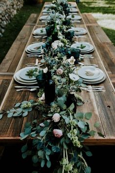 a long wooden table with plates and flowers on it is set for an outdoor dinner