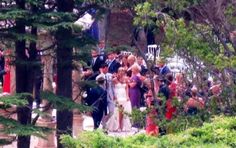 a group of people standing around each other in front of some trees and bushes on a sunny day