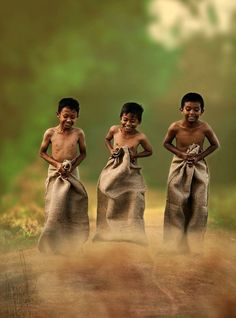 three young boys standing next to each other on a dirt road with grass and trees in the background