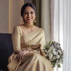 a woman sitting on a couch wearing a gold sari and holding a bridal bouquet