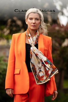 A woman in an orange suit with a patterned scarf, outdoors, looks confidently at the camera.