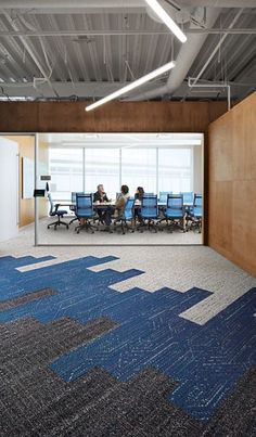 two people sitting at a table in an office with blue chairs and large windows on the wall