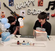 two people sitting at a table with a birthday cake in front of them and balloons on the wall