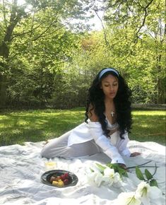 a woman sitting on top of a blanket in the grass next to flowers and fruit