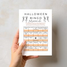 a hand holding up a halloween bingo game on top of a white table with an orange stripe