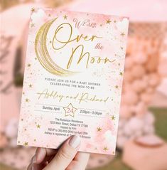 a hand holding up a pink and gold over the moon baby shower card with stars on it