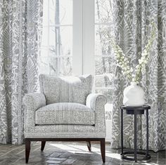 a white chair sitting in front of a window next to a vase with flowers on it